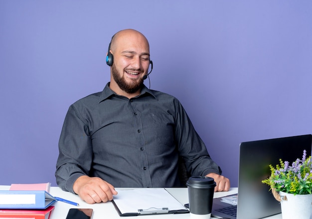 紫色の背景で隔離のラップトップを見て作業ツールと机に座っているヘッドセットを身に着けている若いハゲのコールセンターの男を笑う