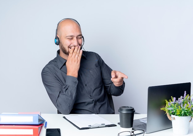 Ridendo giovane calvo call center uomo che indossa la cuffia seduto alla scrivania con strumenti di lavoro guardando e indicando il laptop con la mano sulla bocca isolati su sfondo bianco