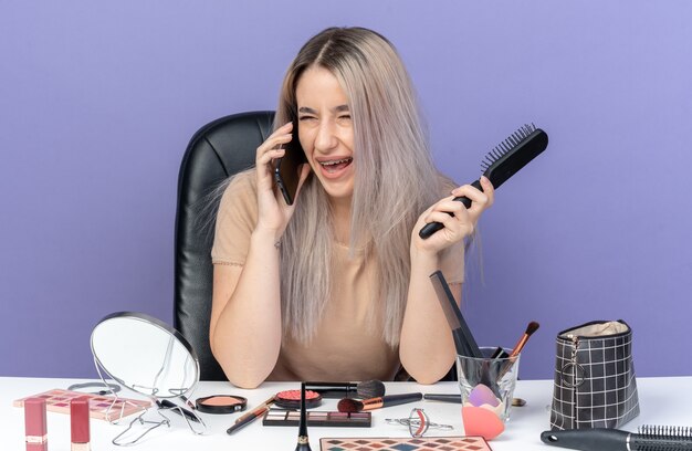 Laughing young beautiful girl wearing dental braces sits at table with makeup tools speaks on phone holding comb isolated on blue wall