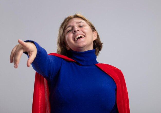 Laughing young blonde superhero woman in red cape looking and pointing at front isolated on white wall