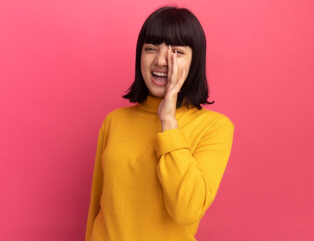 Laughing young brunette caucasian girl holds hand close to mouth on pink 