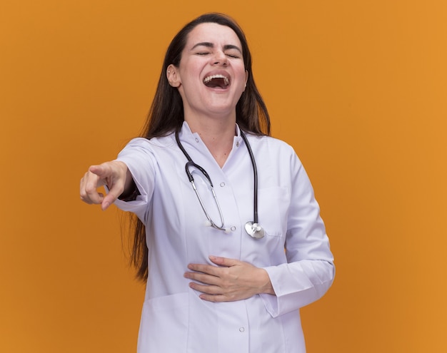 Смеющаяся молодая женщина-врач в медицинском халате со стетоскопом кладет руку на живот и указывает вперед, изолированную на оранжевой стене с копией пространства