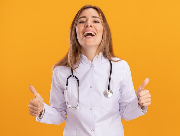 Laughing young female doctor wearing medical robe with stethoscope showing thumbs up isolated on yellow wall