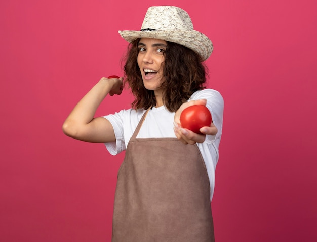 분홍색에 고립 된 토마토를 들고 원예 모자를 쓰고 제복을 입은 젊은 여성 정원사를 웃고