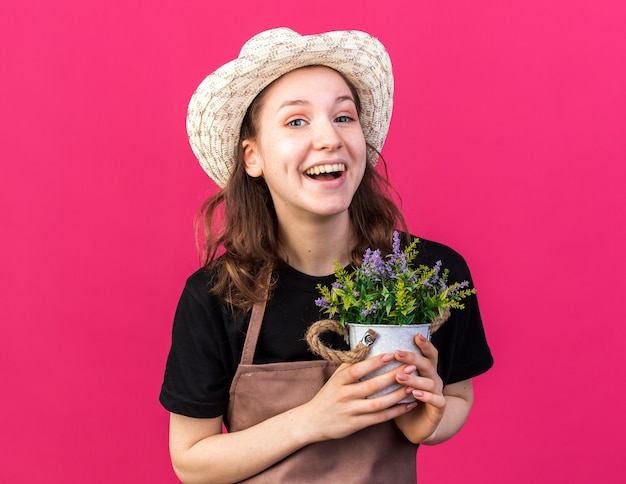 Laughing young female gardener wearing gardening hat holding flower in flowerpot 