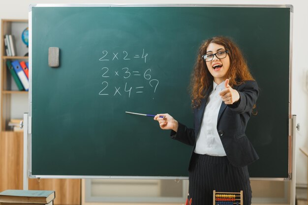 laughing young female teacher standing in front blackboard points at blackboard with pointer in classroom