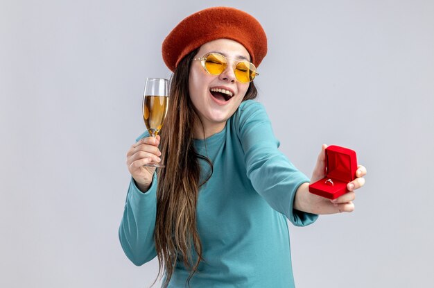 Laughing young girl on valentines day wearing hat with glasses holding glass of champagne with wedding ring isolated on white background