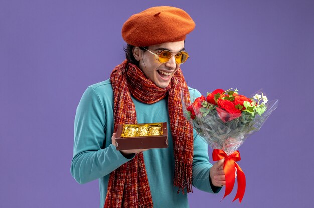 Laughing young guy on valentines day wearing hat with scarf and glasses holding and looking at bouquet with box of candies isolated on blue background