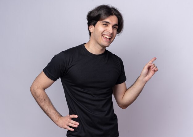 Laughing young handsome guy wearing black t-shirt points at behind putting hand on hip isolated on white wall with copy space