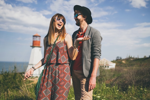 Laughing young hipster couple indie style in love walking in countryside, lighthouse on background