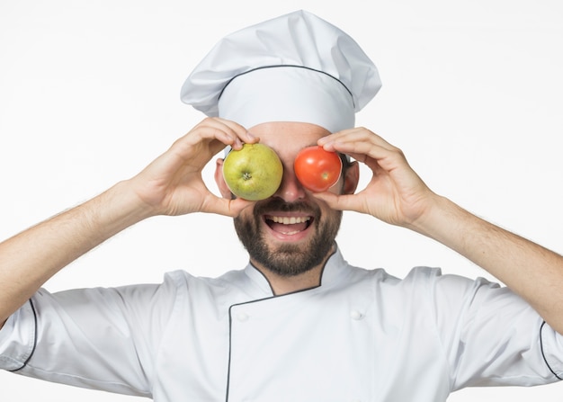 Free photo laughing young male chef holding apple and red tomato in front of his eyes