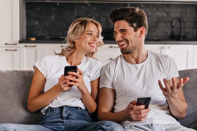 Laughing young man talking with wife on sofa. Pretty girl chilling on couch and texting message.