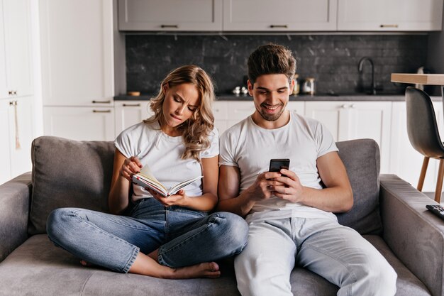 Laughing young man texting message. Serious woman reading book while sitting on couch with husband.