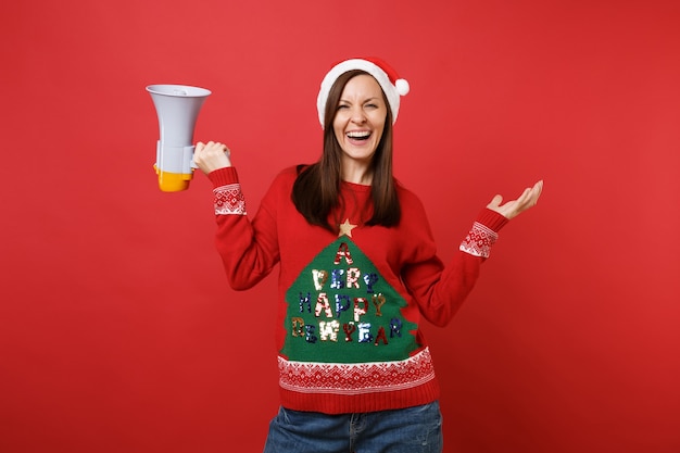 니트 스웨터를 입은 어린 산타 소녀, 확성기를 들고 크리스마스 모자를 쓰고 빨간 배경에 고립된 손을 펼치고 있습니다. 새해 복 많이 받으세요 2019 축 하 휴일 파티 개념입니다. 복사 공간을 비웃습니다.