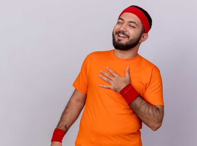 Laughing young sporty man with closed eyes wearing headband and wristband putting hand on chest