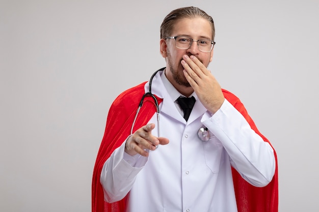 聴診器と眼鏡で医療ローブを着て笑っている若いスーパーヒーローの男は、白い背景で隔離されたジェスチャーを示す手で口を覆った