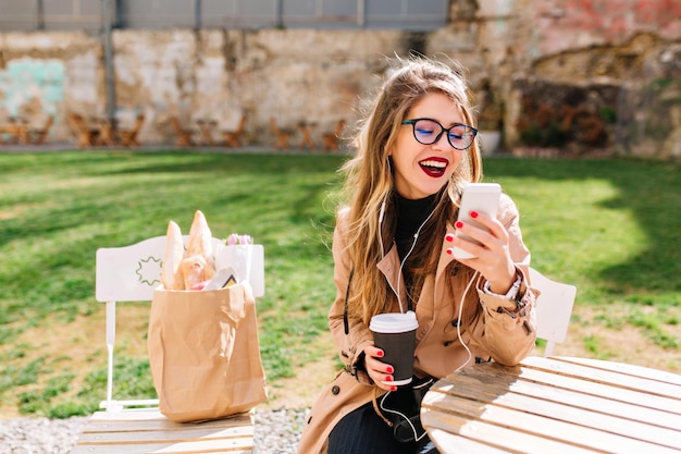 Laughing young woman in glasses rests in outdoor cafe after a walk and listens to music on the phone. Hipster girl checks the mail on social networks sitting in the park after shopping. Using devices