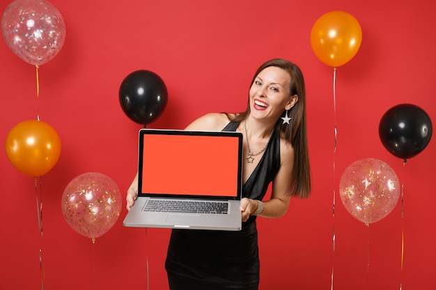 밝은 빨간색 배경 공기 풍선에 빈 검은색 빈 화면이 있는 노트북 컴퓨터를 들고 축하하는 검은 드레스를 입은 젊은 여성을 웃고 있습니다. 새해 복 많이 받으세요, 생일 모의 휴일 파티 컨셉입니다.