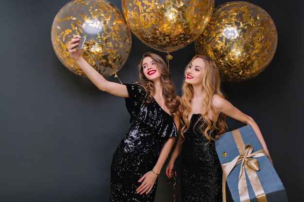 Laughing young woman with curly hairstyle posing with pleasure during party. Glamorous birthday girl in black outfit holding big gift box while her friend making selfie.