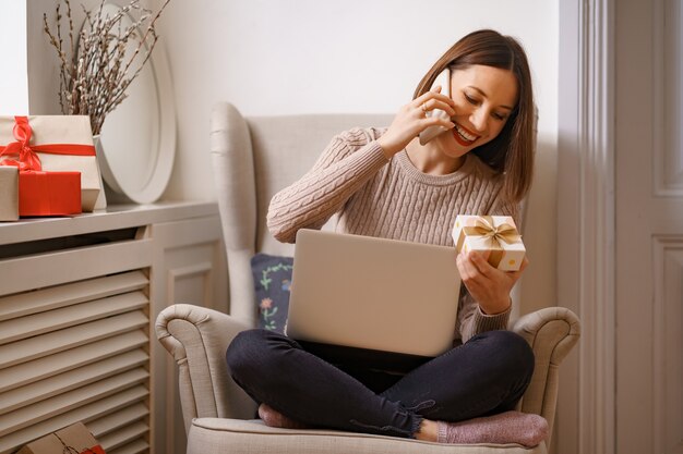 快適な肘掛け椅子に座って携帯電話で話しているラップトップで笑う若い女性