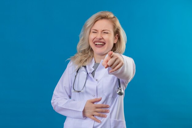Laughting young doctor wearing stethoscope in medical gown points to camera on blue wall