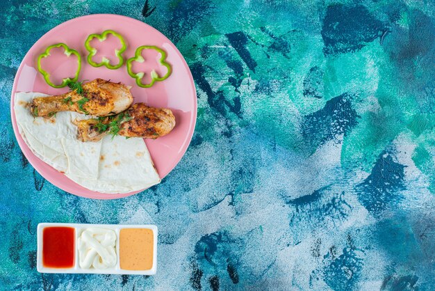 Lavash, baked drumsticks and pepper on a plate next to sauce bowls close up , on the blue background. 