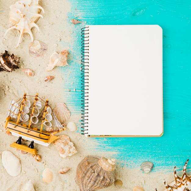 Layout of seashells and toy ship among sand near notebook