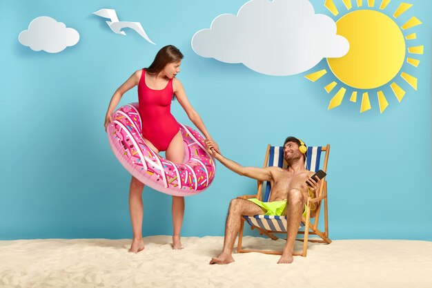 Lazy guy sits at beach chair, enjoys listening pleasant music, stretches hand to girlfriend who stands in pink inflatable swimming ring
