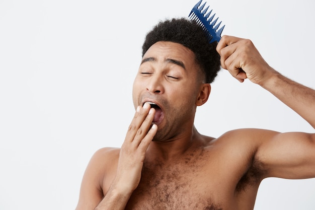 Mattina pigra. ritratto di bello maschio afroamericano divertente con capelli ricci senza vestiti vestiti bocca, sbadigliando cercando di pettinare i capelli in mattinata.