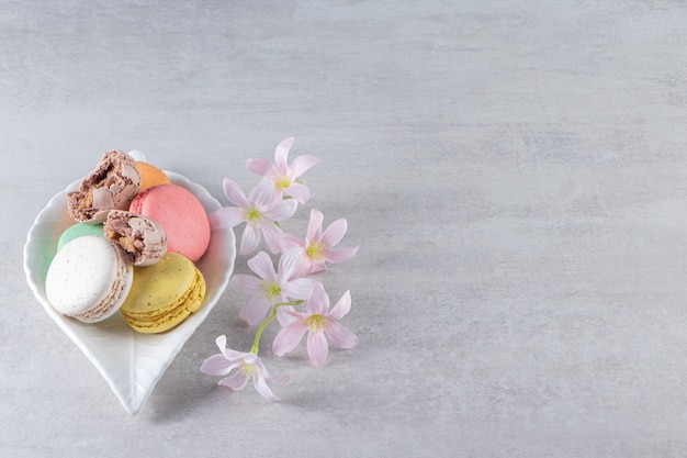 Leaf-shaped plate of colorful sweet macaroons with flowers on stone table. 