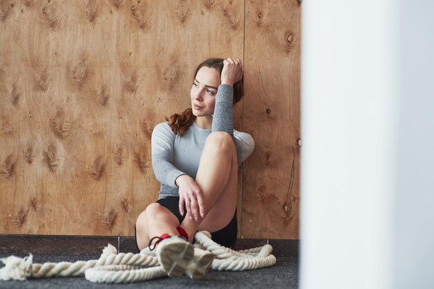Опираясь на деревянную стену. Спортивная молодая женщина имеет фитнес-день в тренажерном зале в утреннее время