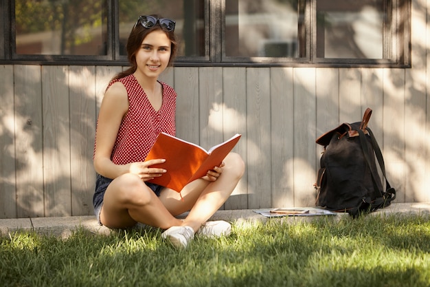 Learning, education, people and lifestyle concept. Summertime image of beautiful student girl sitting on green grass in park, keeping legs crossed, holding copybook with lectures, preparing for exam