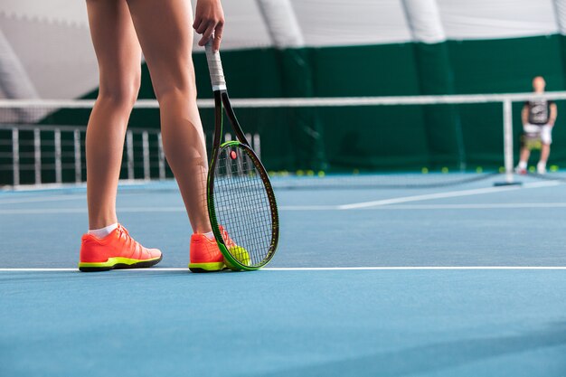 Ноги молодой девушки в закрытом теннисном корте с мячом и ракеткой