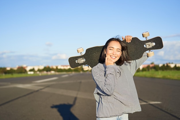 田舎の空の道をクルージングするロングボードを持って立つレジャーと人々の幸せなアジア人女性