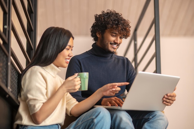 Leisure. Young long haired asian woman with cup sideways to camera pointing at screen and curly bearded indian man cheerful looking at laptop sitting on stairs at home