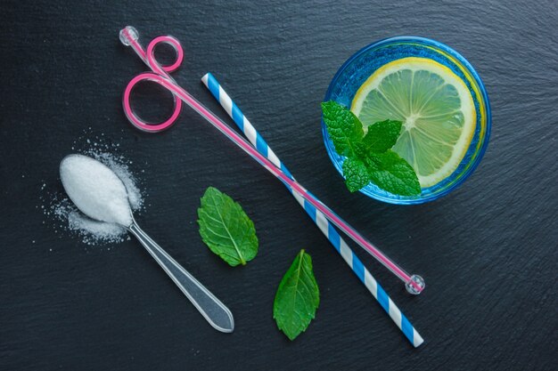 Lemon slice in a blue bowl with straws, leaves and spoon with salt top view on a dark stone surface