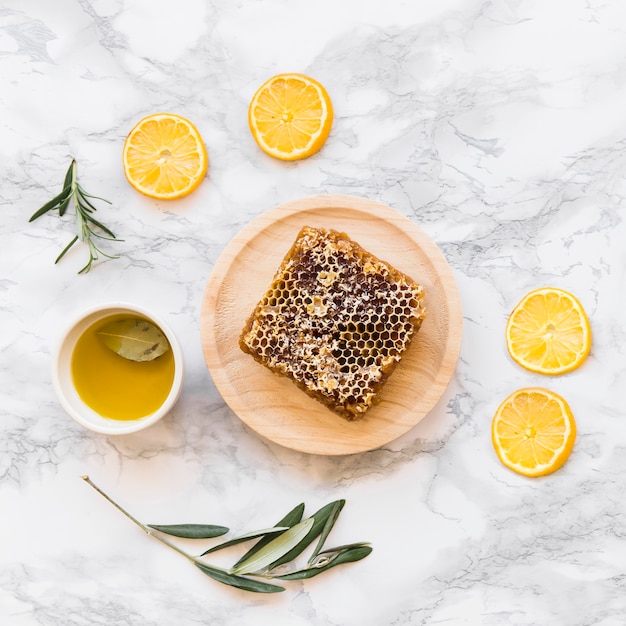 Lemon slice, twig with honeycomb and bowl of oil on white marble background