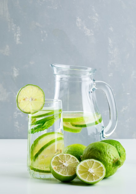 Lemonade in glass and jug with lemon, basil side view on white and plaster