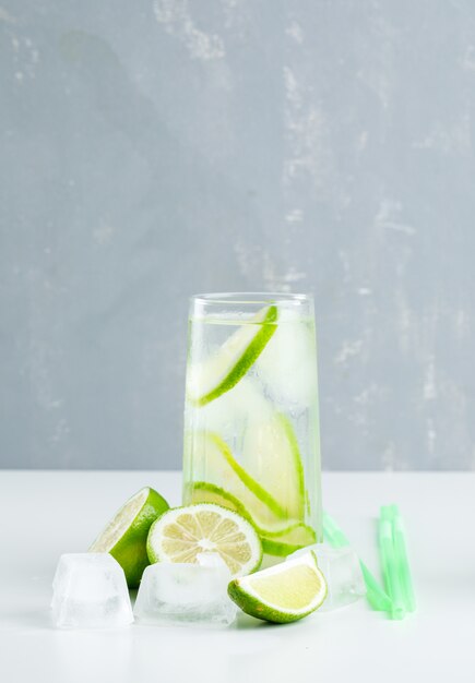 Lemonade in a glass with lemon, straws, ice cubes side view on white and plaster