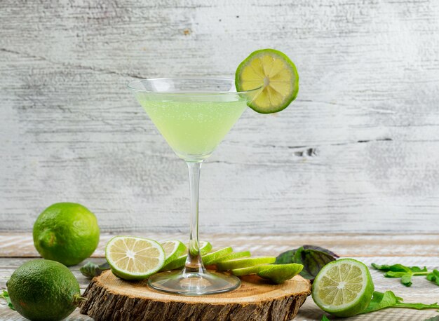 Lemonade in a glass with lemons, leaves, cutting board side view on wooden and grungy