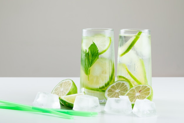 Lemonade in glasses with lemons, straws, basil, ice cubes side view on white and grey