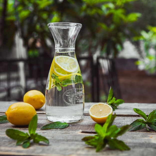 Lemonade and ingredients in a glass jug on wooden and garden table, side view.