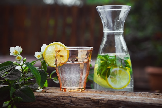 Lemonade with flowers on branch in glass and jug on wooden and yard table, side view.