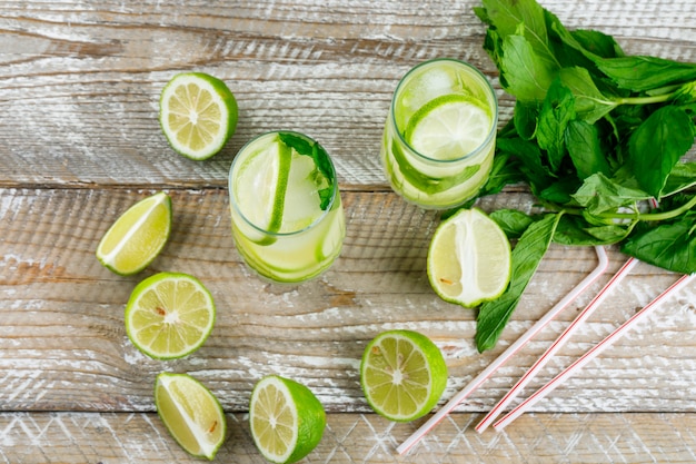 Lemonade with lemon, basil, straws in glasses on wooden, flat lay.