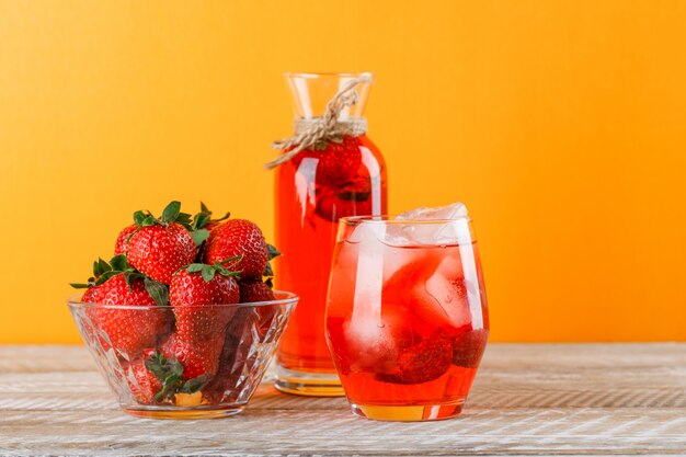 Lemonade with strawberries in jug and glass on wooden and yellow background, side view.