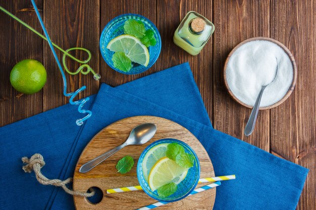 Lemons in a bowl with blue cloth, wooden knife and bottle of juice, straws, bowl of salt top view on a wooden surface