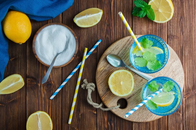 Лимоны в миске с синей тканью, деревянным ножом и бутылкой сока, соломкой, миской с солью, вид сверху на деревянной поверхности