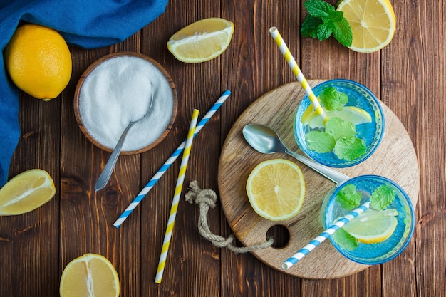 Lemons in a bowl with blue cloth, wooden knife and bottle of juice, straws, bowl of salt top view on a wooden surface