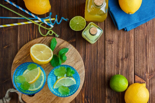 Lemons in a bowl with blue cloth, wooden knife and bottle of juice, straws, leaves top view on a wooden surface