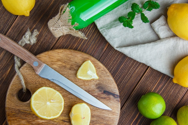 Lemons with limes, leaves, knife, drink, cutting board on wooden and kitchen towel, flat lay.